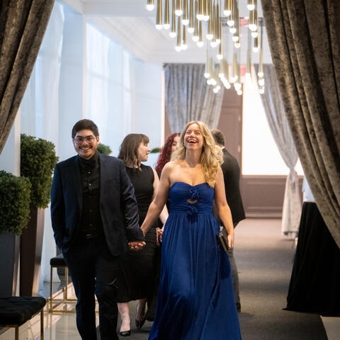 Des participants arrivent à un gala en marchant sous des ballons noir et or et des lumières suspendues chaleureuses à l’entrée.