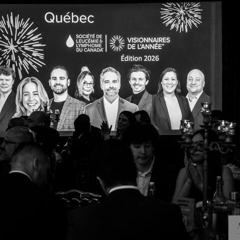 Des participant·e·s observent et applaudissent tandis qu’un grand écran met en valeur des personnes honorées lors d’un gala-bénéfice en salle sombre.